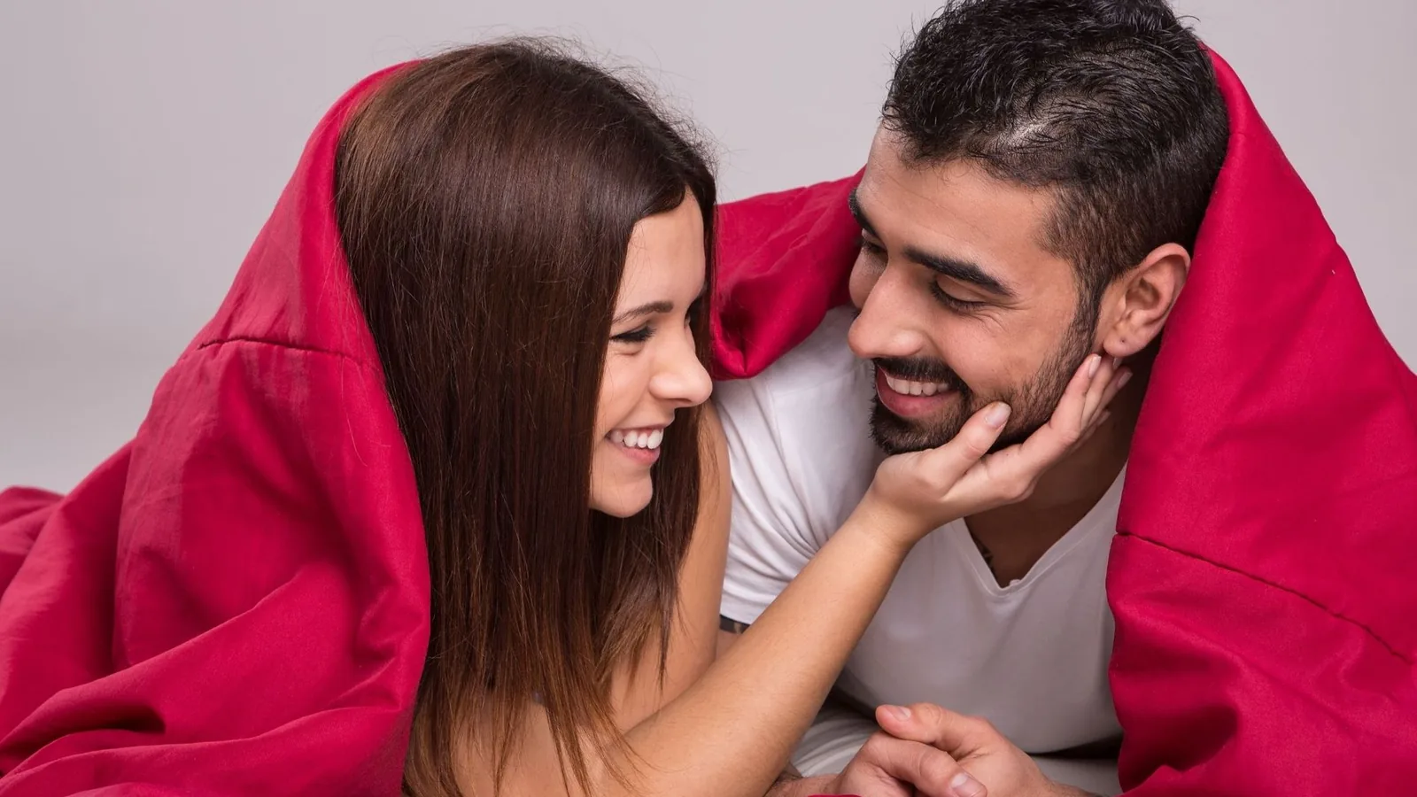 A smiling couple lies facing each other on a bed with a red blanket pulled over both their heads, her hand touching his beard