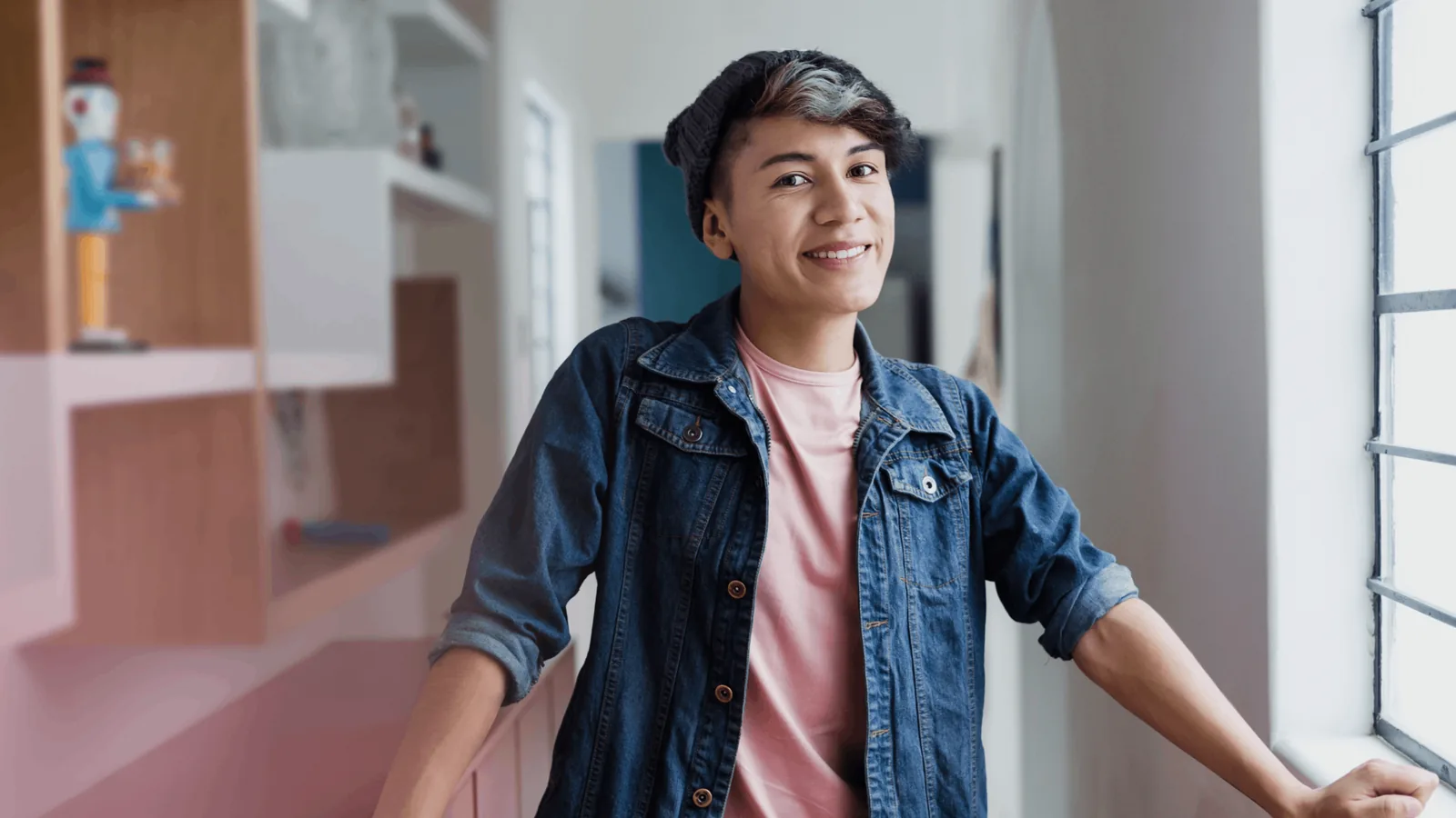 A smiling young person in a denim jacket leaning by a window at home
