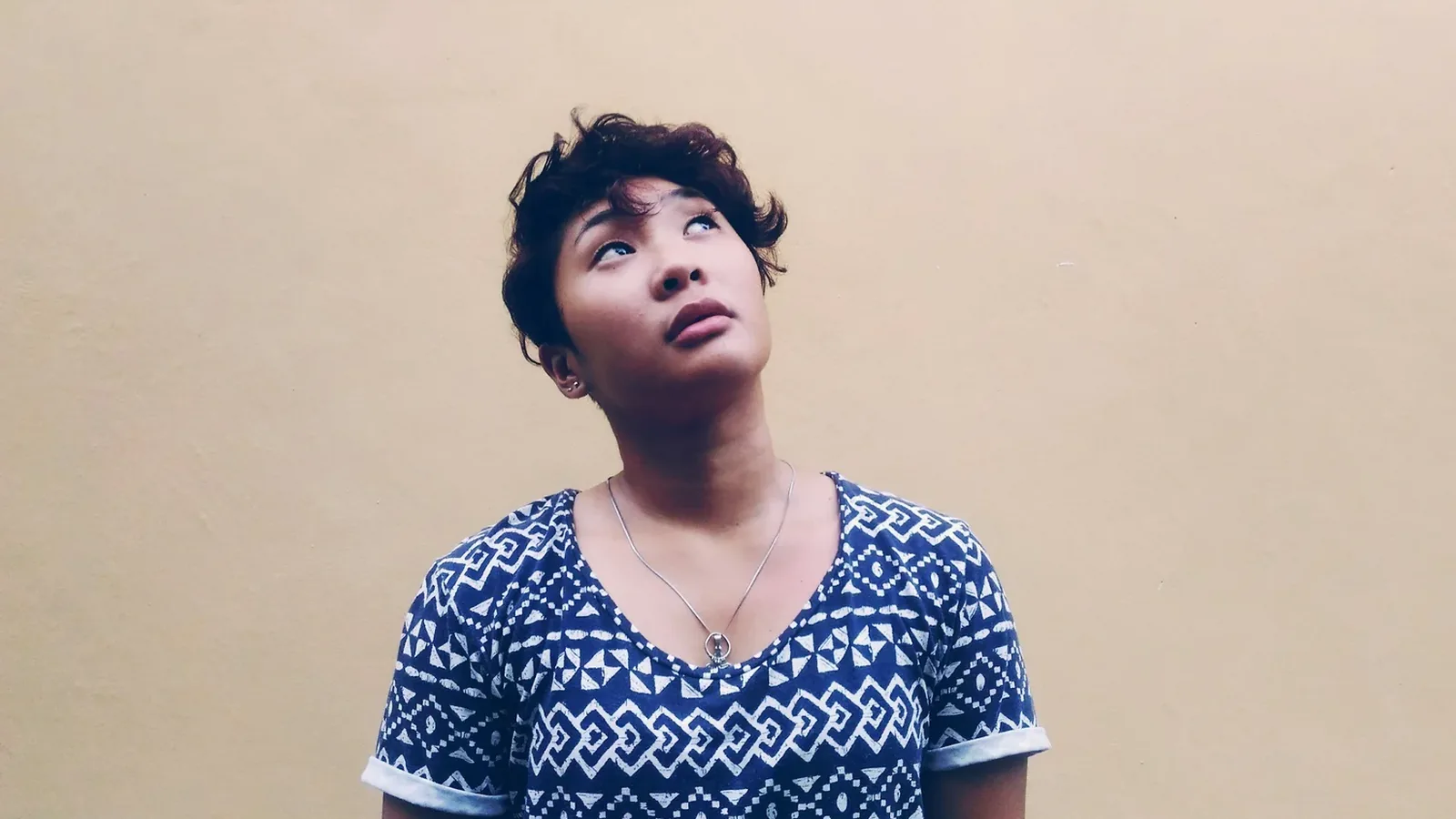 A young person in a patterned T-shirt looking up thoughtfully against a beige wall