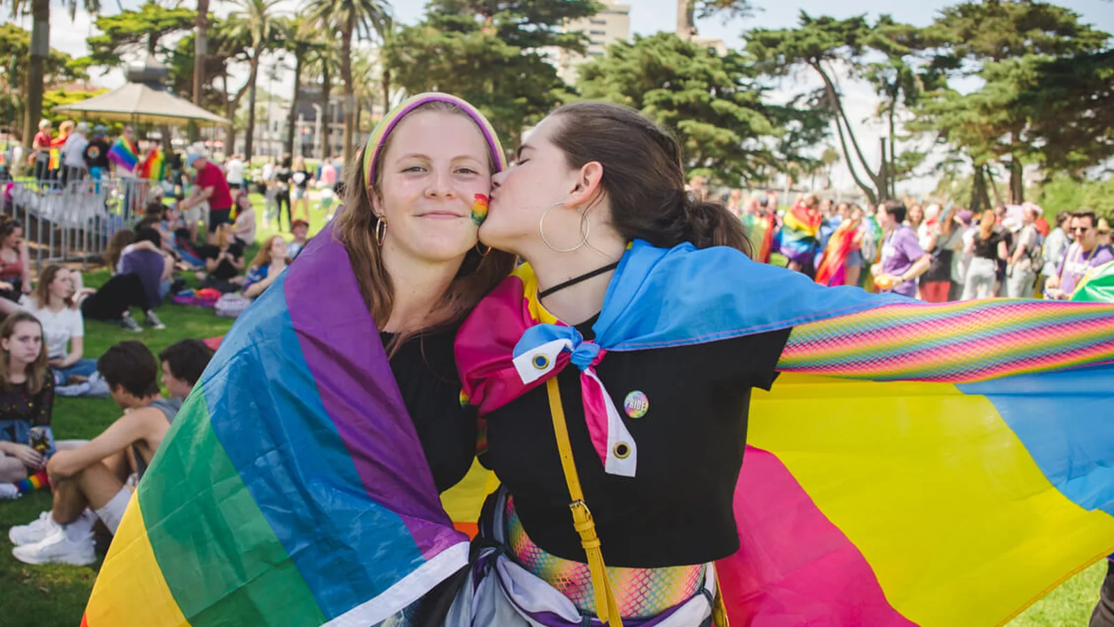 Two women wrapped in pride flags at a park festival, one kissing the other's cheek