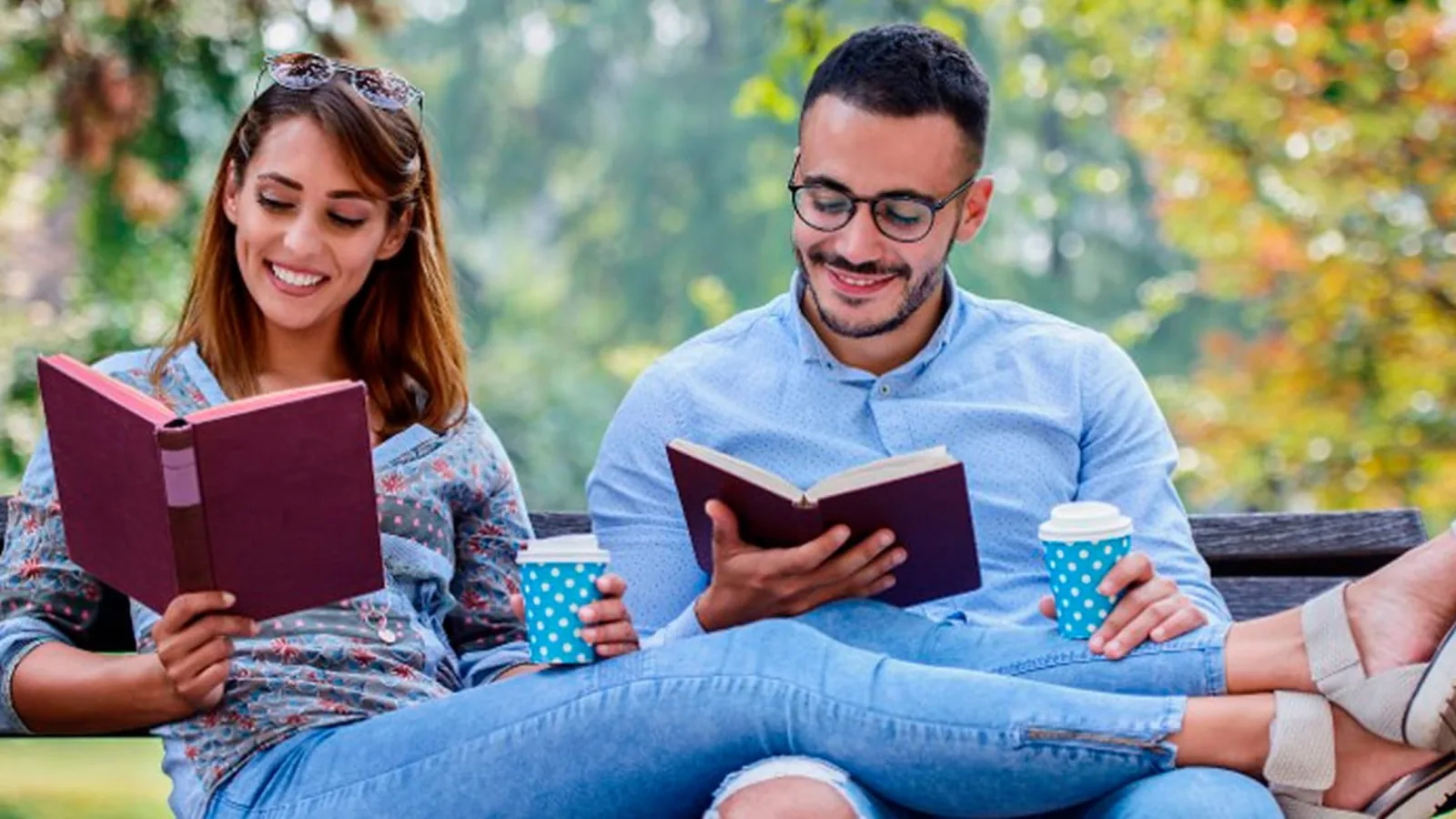 A smiling couple reading books side by side on a park bench