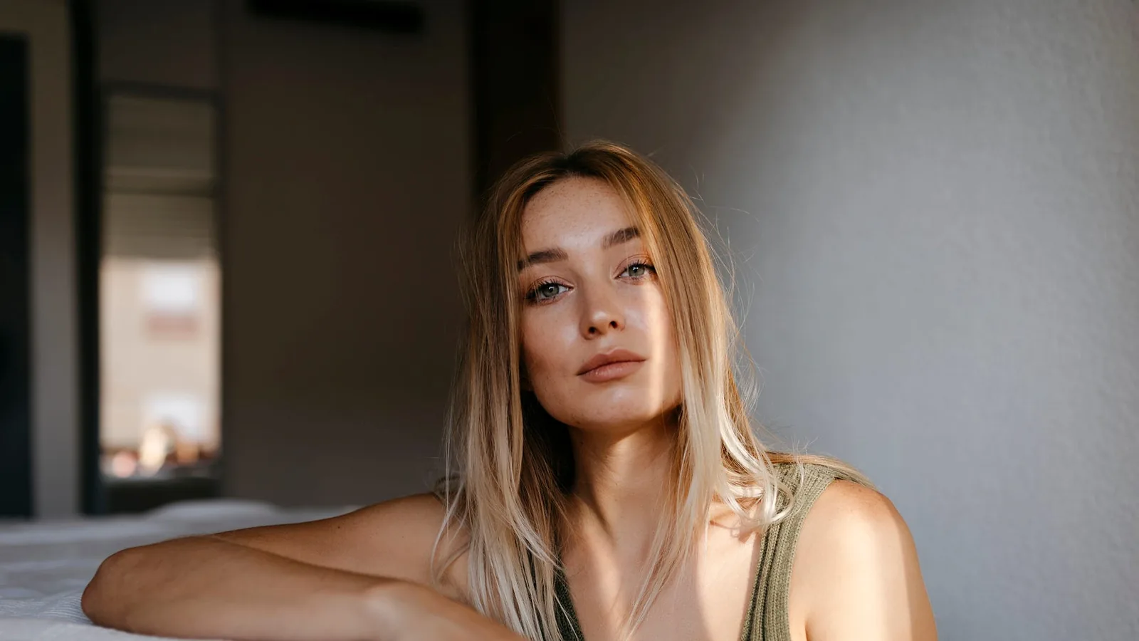 A woman in an olive sleeveless top leaning on the edge of a bed in a bright bedroom, looking toward the camera in soft window light