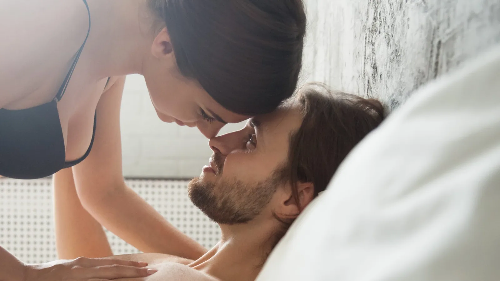 A couple lying together on a bed in warm light, the woman in underwear leaning over the shirtless man as they kiss