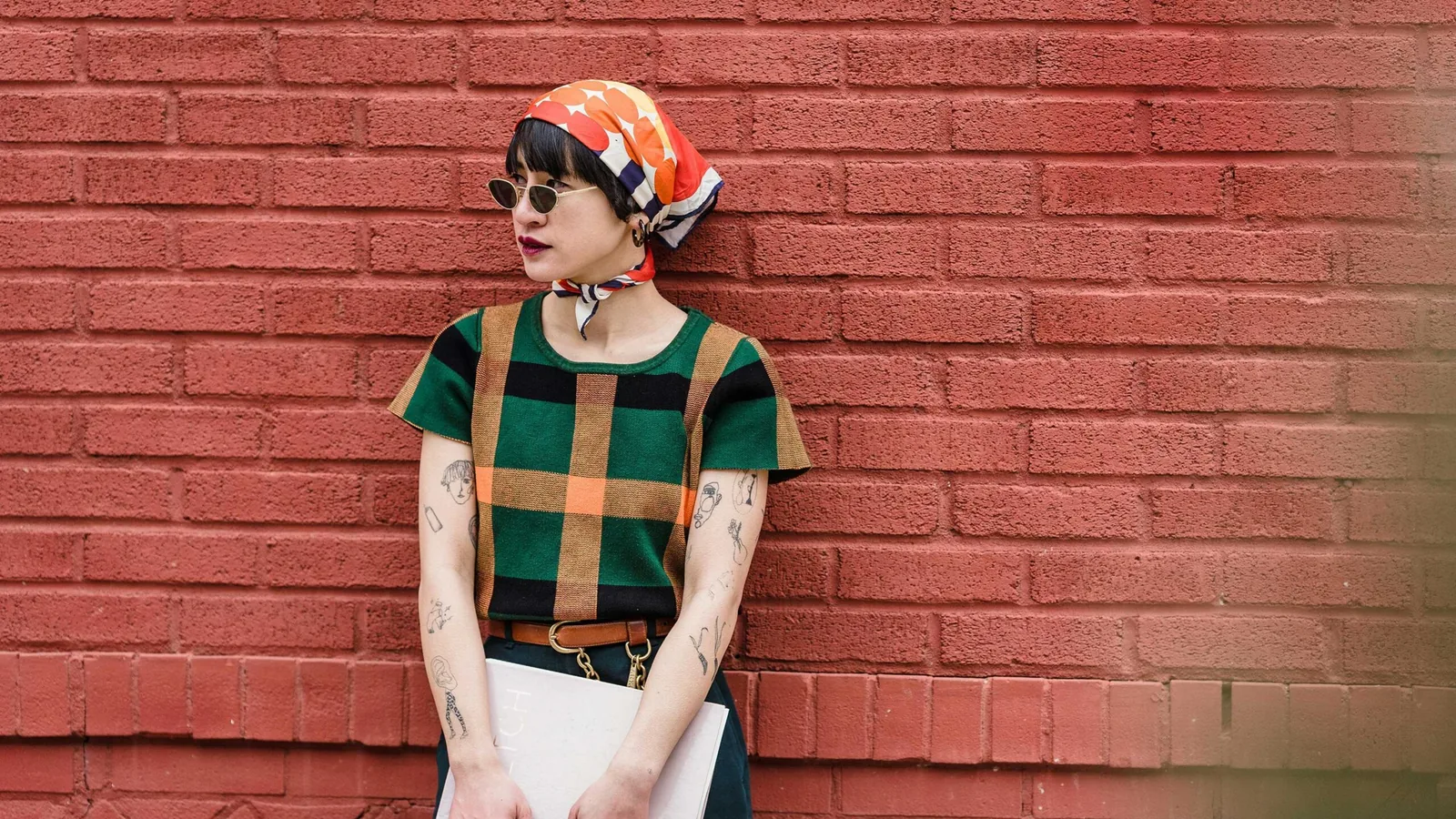 A young woman in a headscarf and sunglasses leaning against a red brick wall