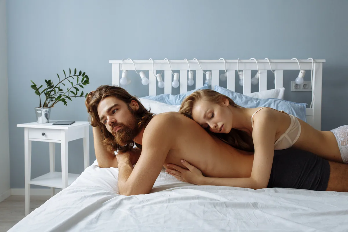 A woman in a bra and underwear rests draped over her partner’s back as he lies face-down on a white bed