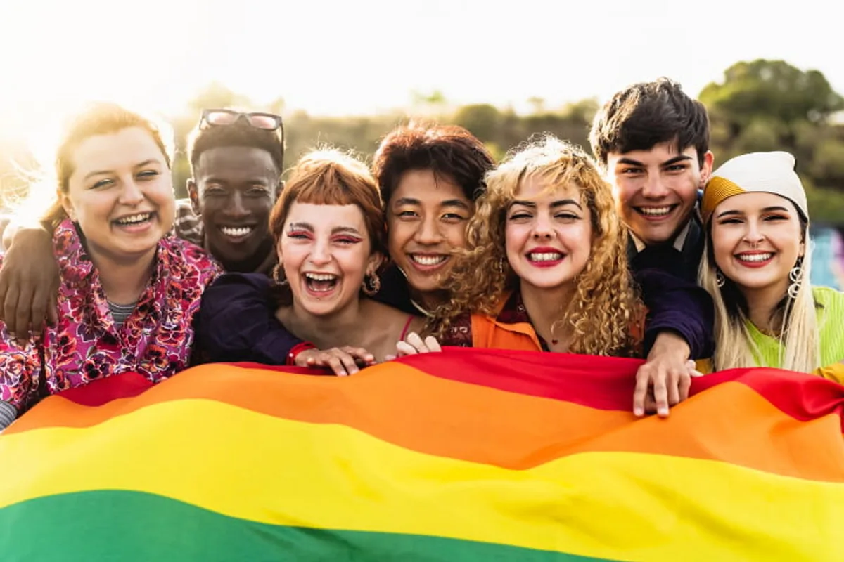 A laughing group of young adults holding a rainbow flag outdoors