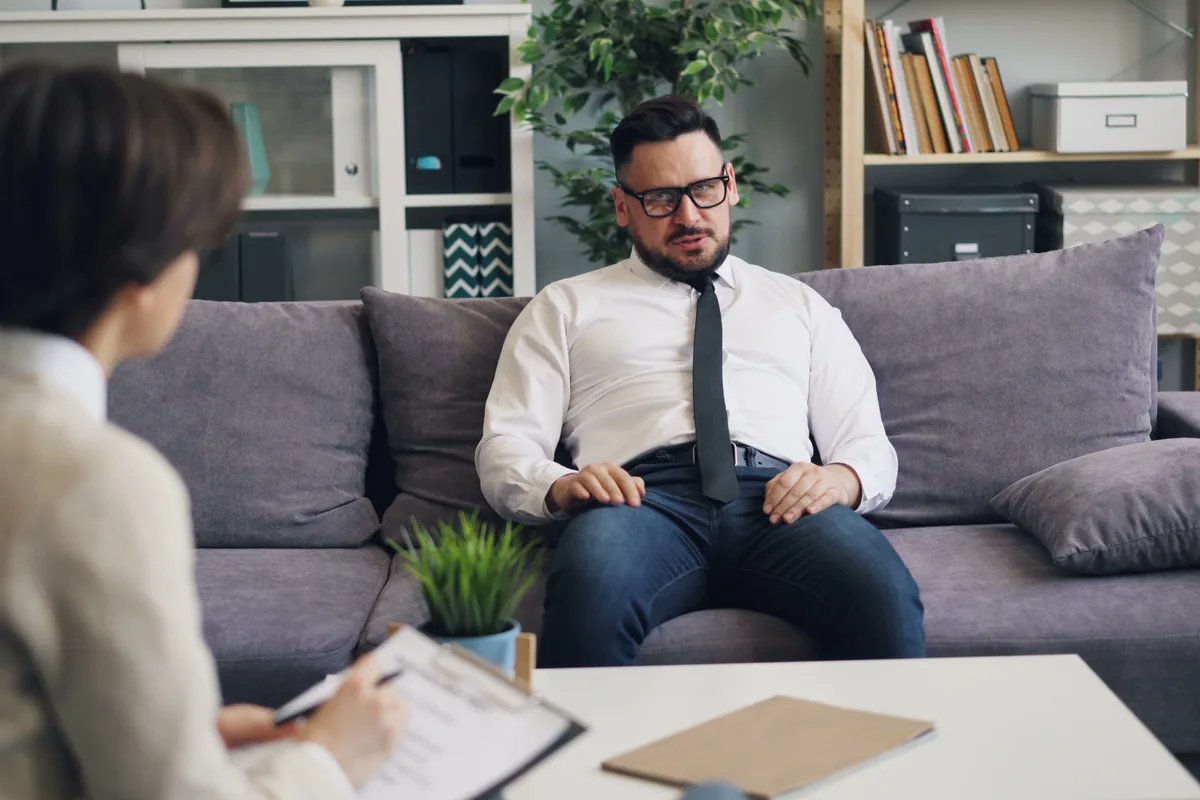 A man on a sofa talking with a therapist who takes notes
