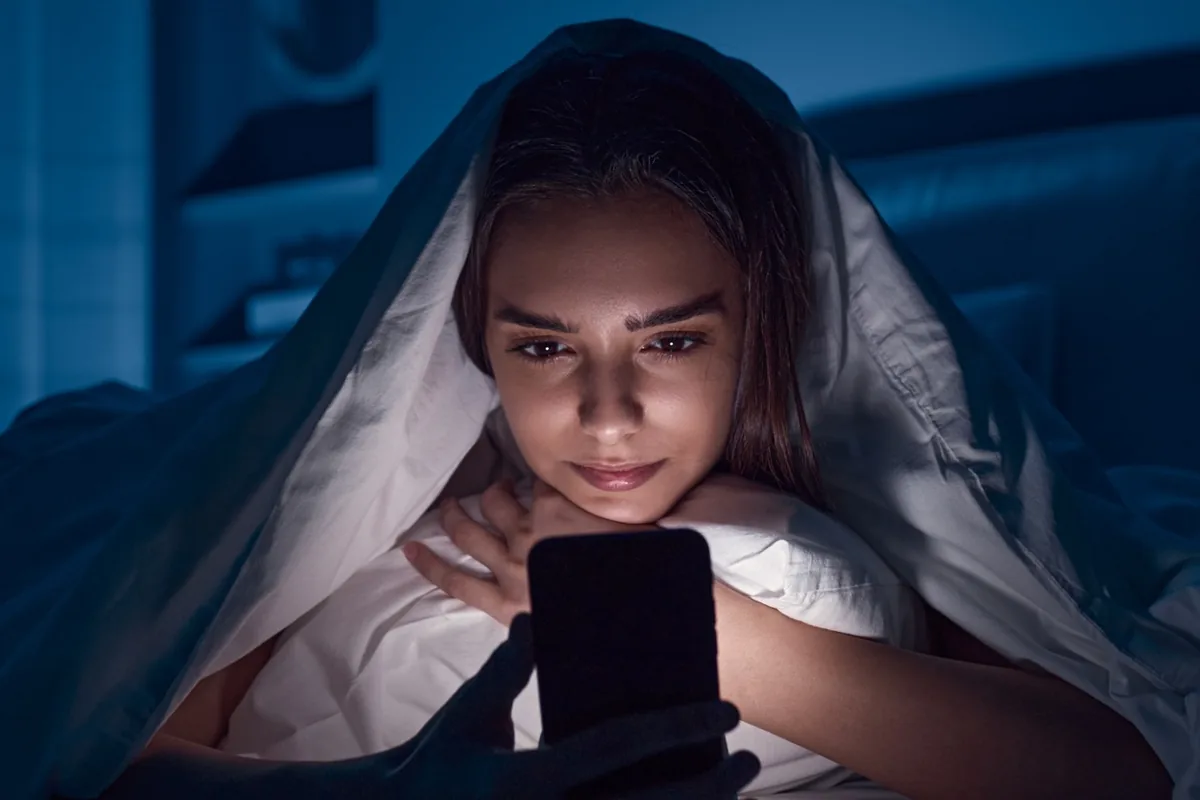 A young woman under a blanket reading her phone at night