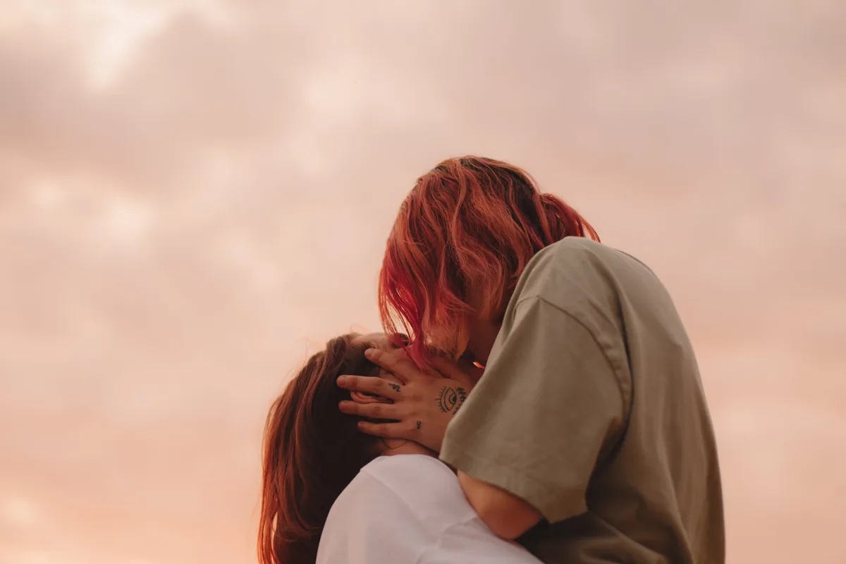 Two people kissing and holding each other against a soft pink evening sky