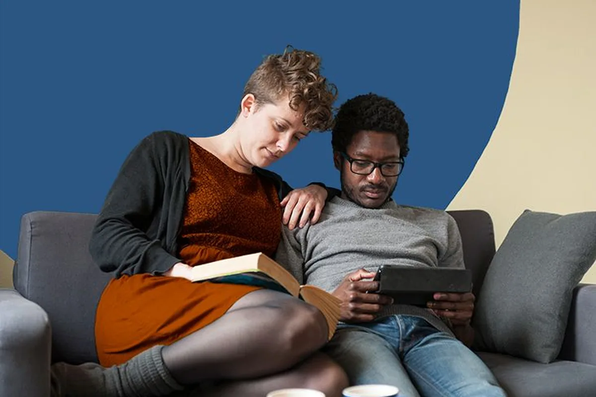 A couple curled up on a sofa, one reading a book and one a tablet