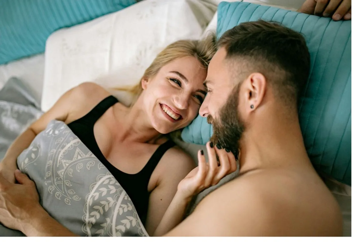 A young couple lying in bed facing each other and smiling warmly, surrounded by teal corduroy pillows and white bedding