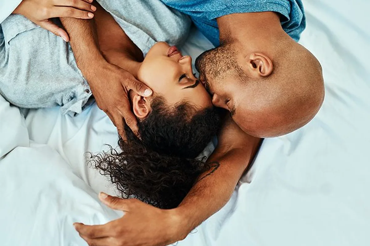 Overhead photo of a partner in a blue shirt tenderly kissing the forehead of a woman with dark curly hair as they lie on white bedding