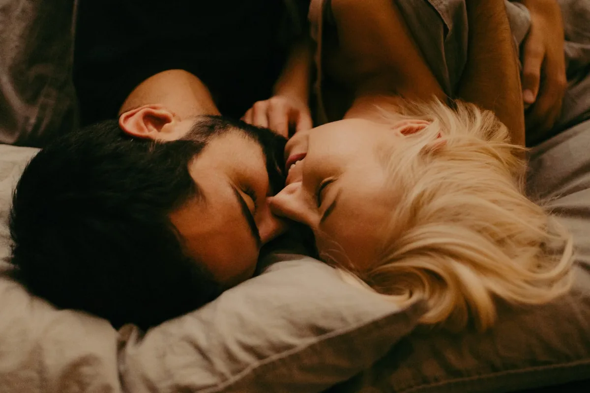 A couple lying in bed embracing in warm lamplight, the smiling woman close to the bearded man’s face