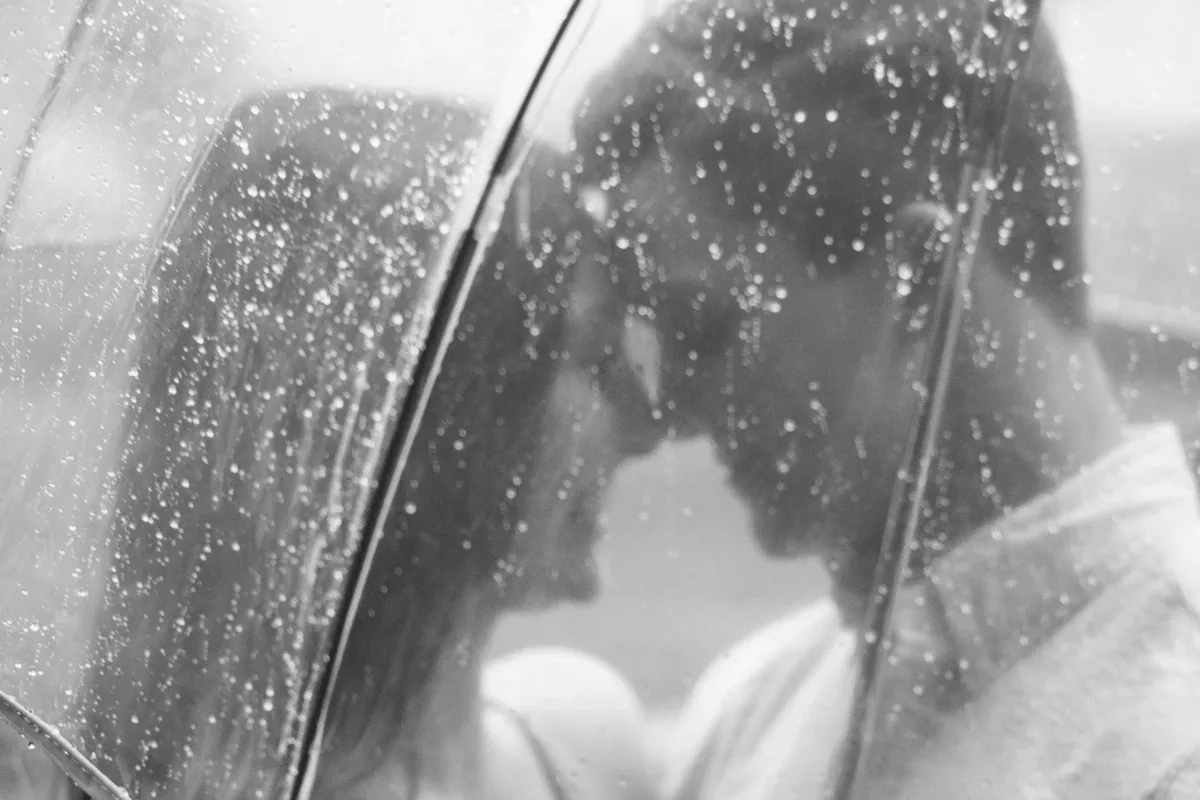 A couple leans toward each other under a clear umbrella in the rain, about to kiss