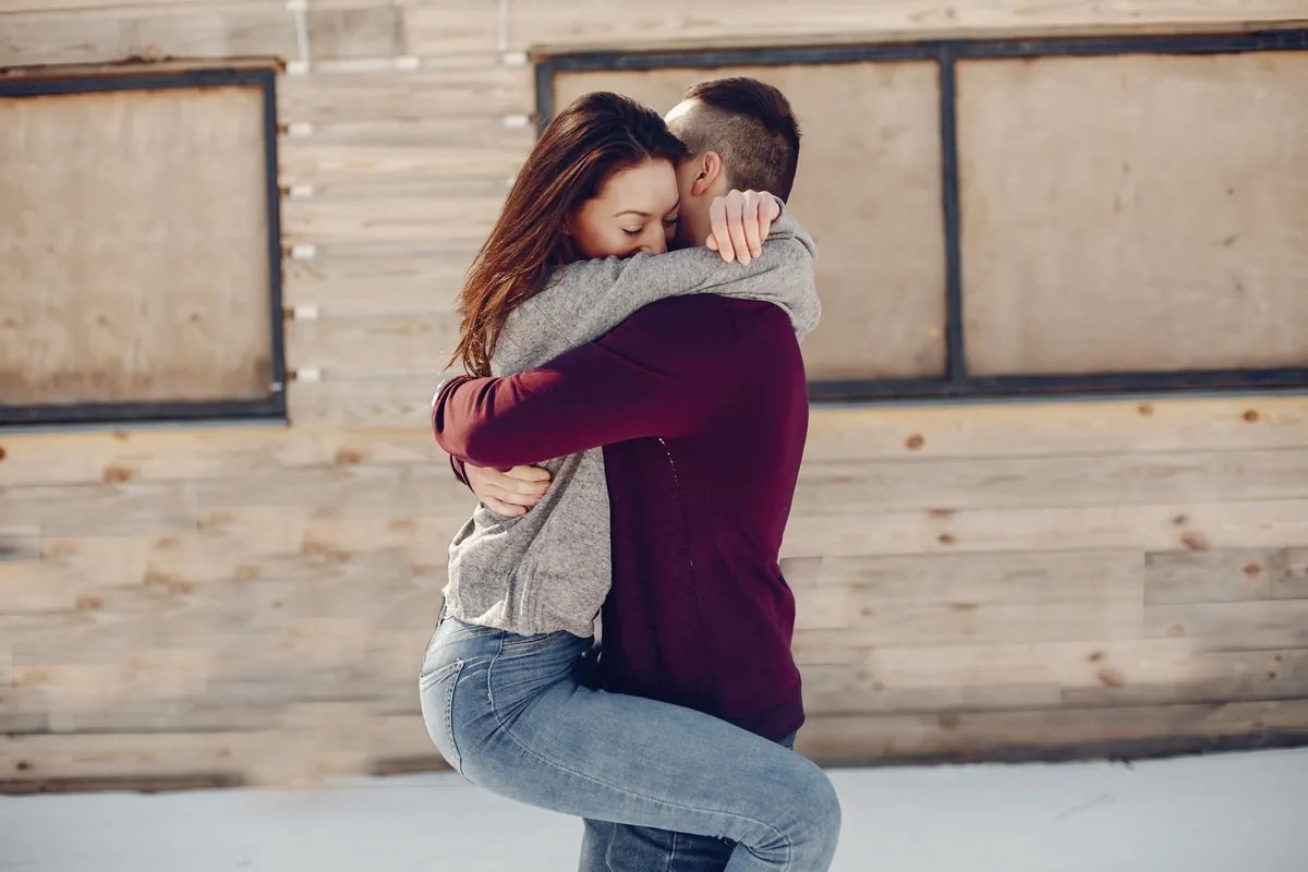A couple in sweaters embracing outdoors in front of a snowy building, the woman wrapping her legs around the man as he lifts her