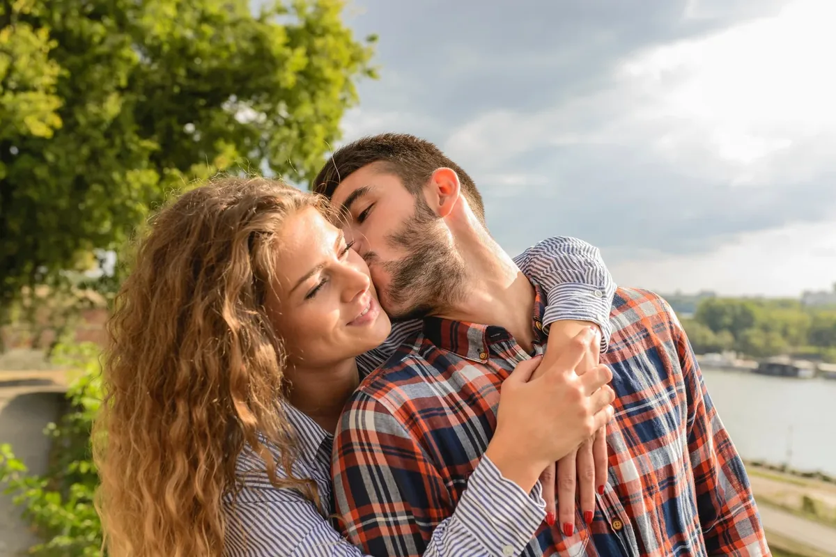 A smiling couple in flannel and striped shirts hugging by a river, the man kissing the woman’s temple