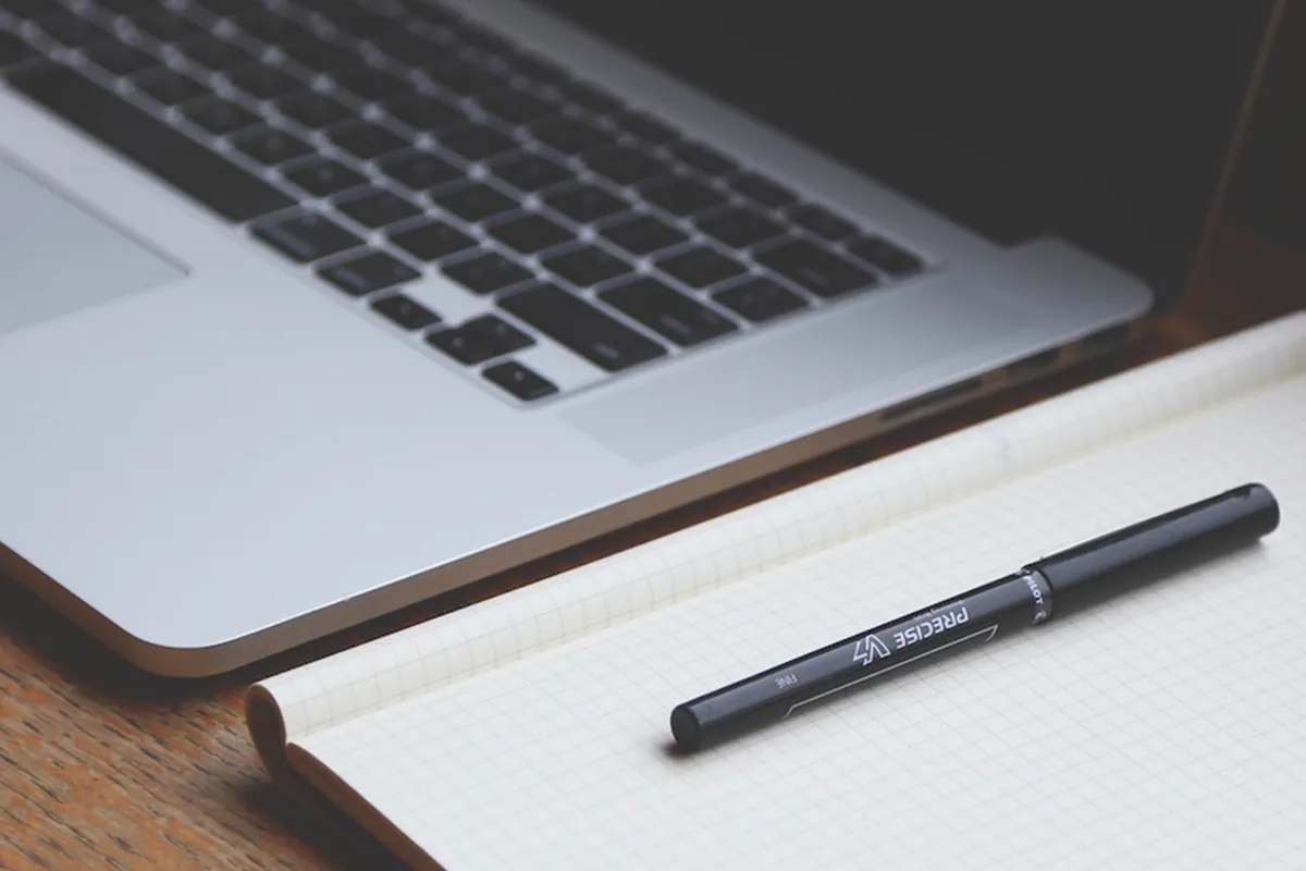 A laptop beside an open notebook and a pen on a wooden desk
