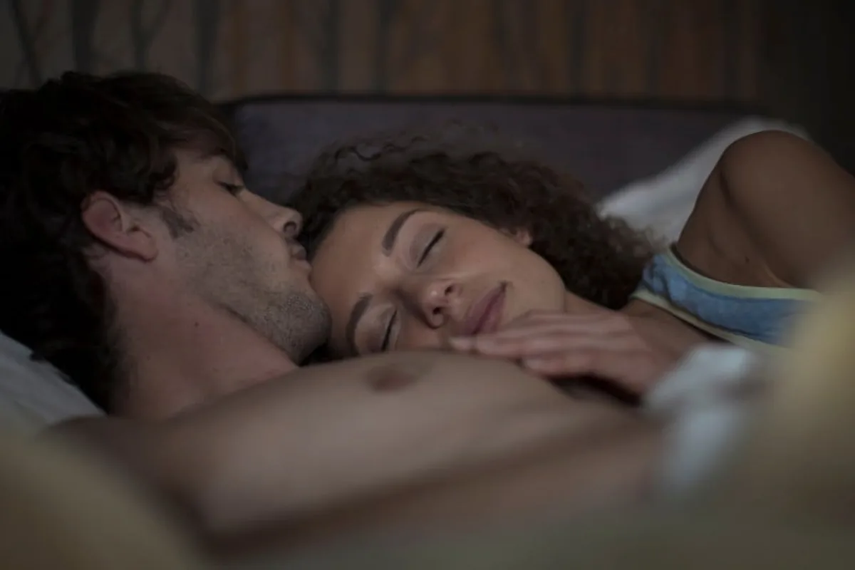 A couple resting in bed in dim warm light, the shirtless man on his back and the woman in a tank top lying against him with her hand near her face