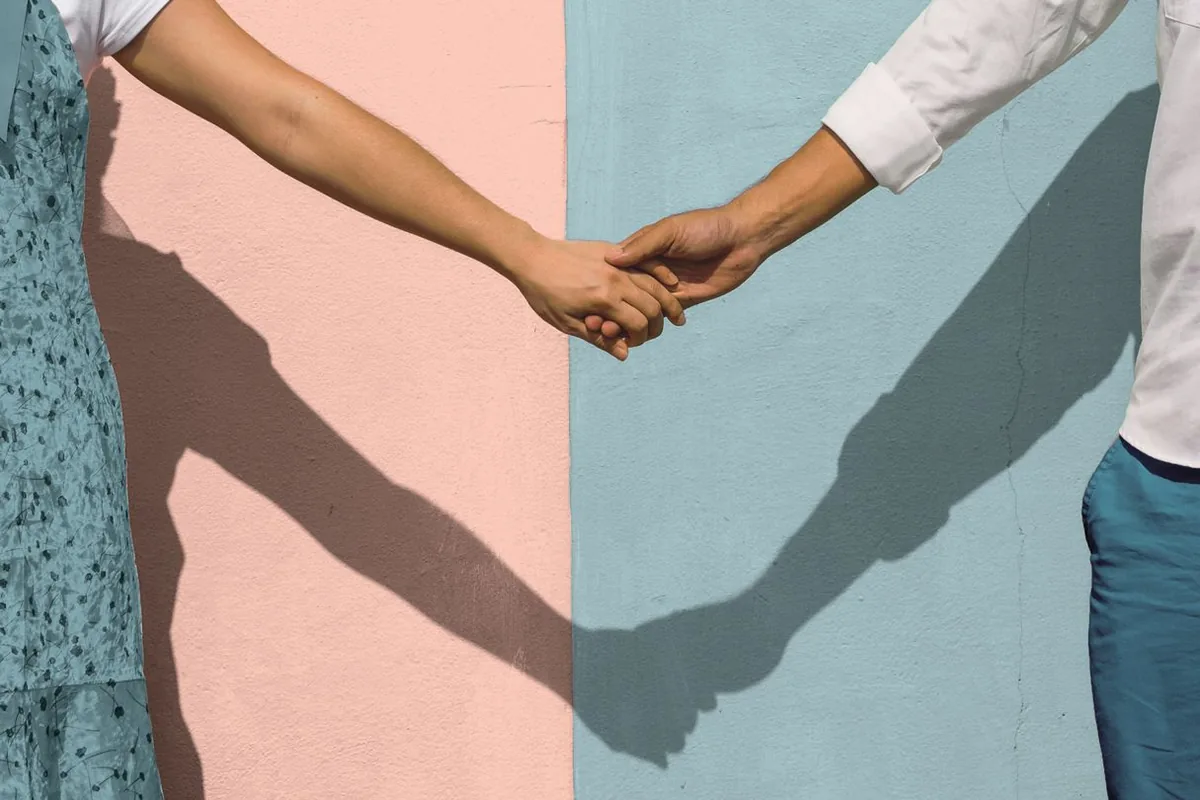 Two people holding hands in bright sunlight against a two-toned pink and blue wall, their faces out of frame