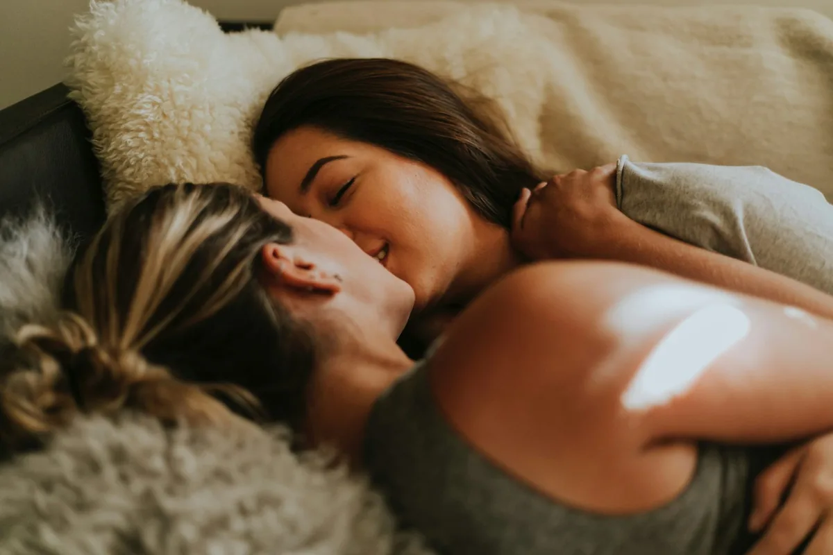 Two clothed women lying close together on a couch under soft blankets, one nuzzling the other affectionately