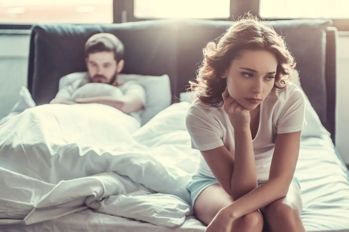 A woman sitting on the bed looking away while her partner lies behind her, both unsettled