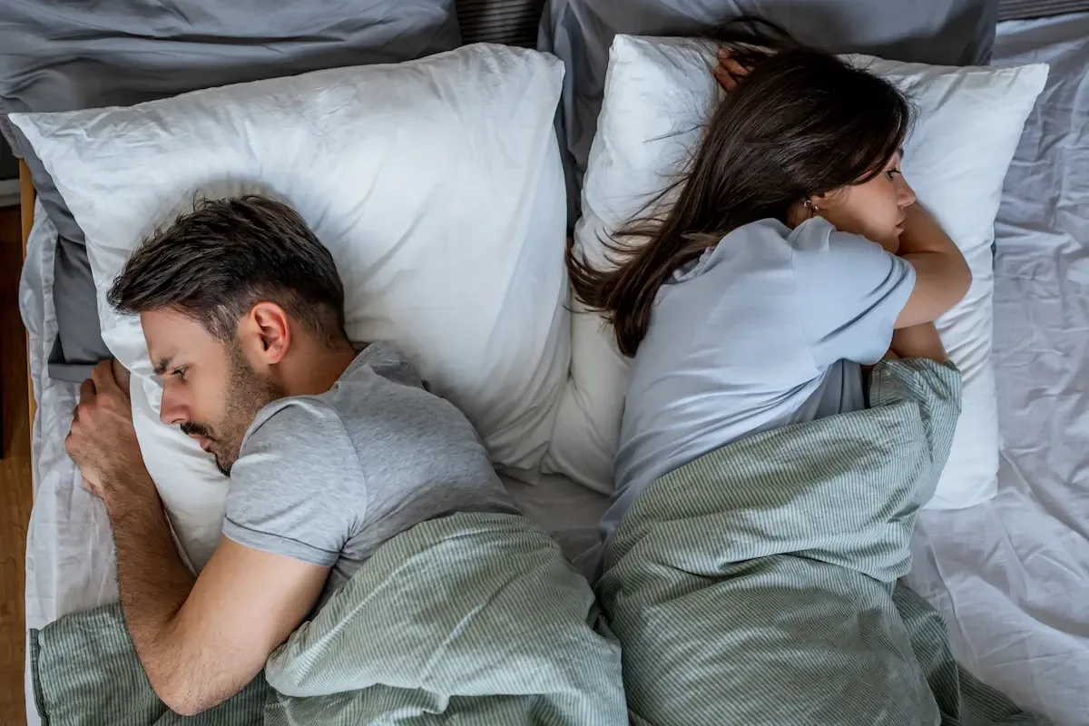 A couple lying back-to-back in bed, both awake and facing away from each other, conveying emotional and physical distance