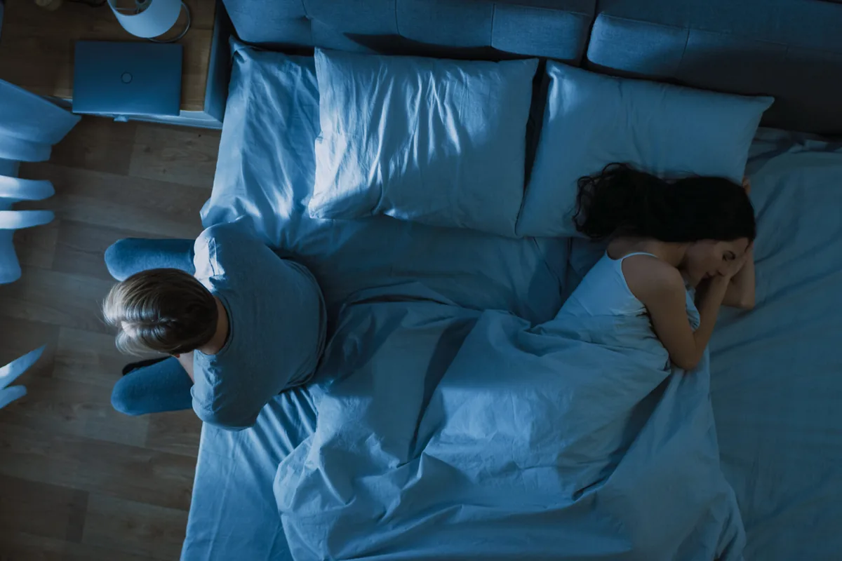 An overhead night-time view of a dim, blue-lit bedroom where a woman sleeps in bed while a man sits on the floor beside it, hunched over with his head bowed