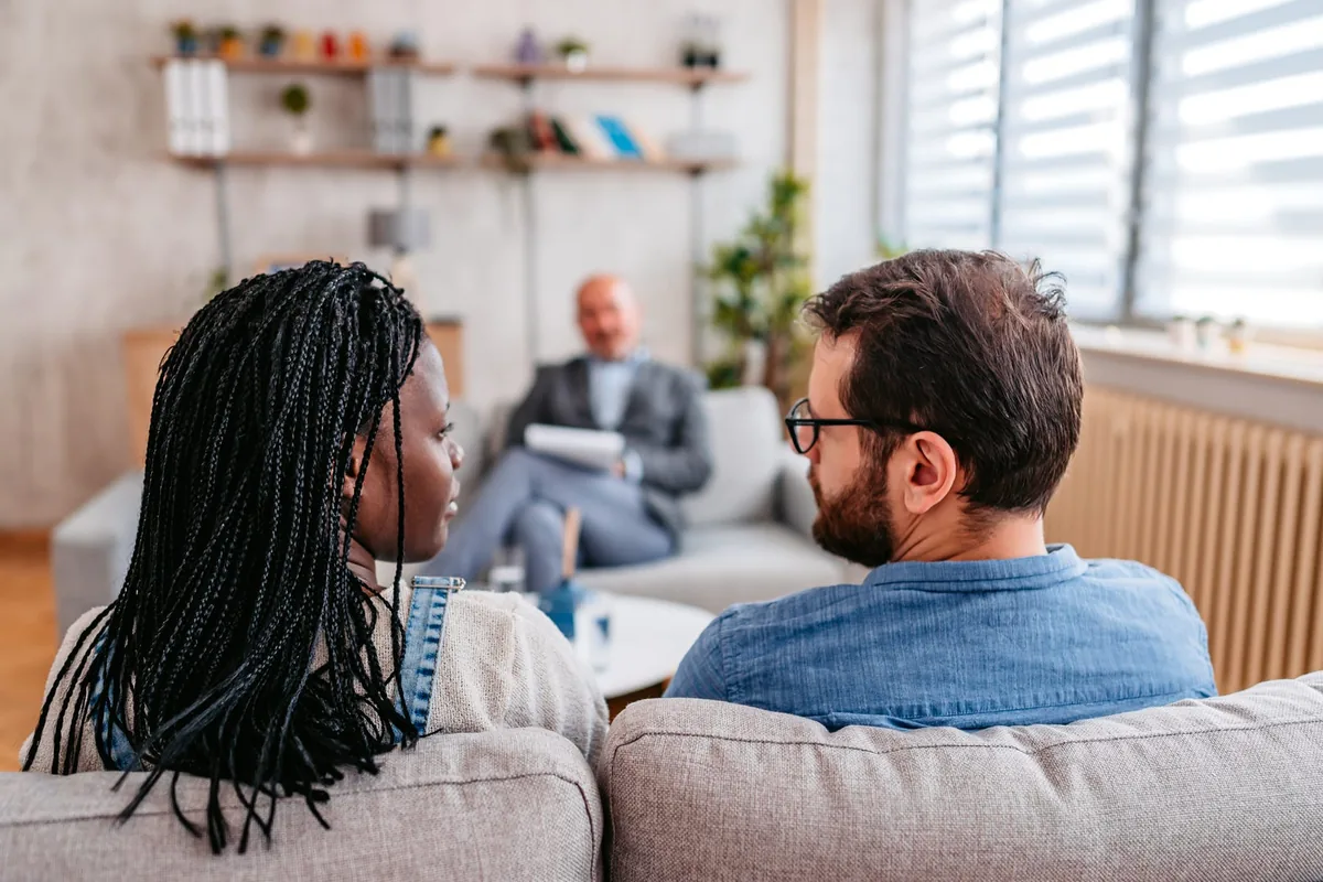 A couple seated together on a sofa in a bright counseling office, viewed from behind as a therapist with a notepad sits across from them
