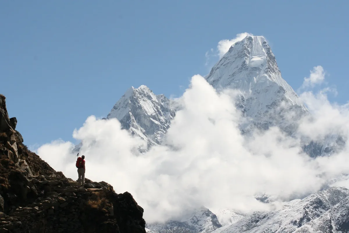 A hiker standing on a ridge below a snowy mountain peak wrapped in clouds