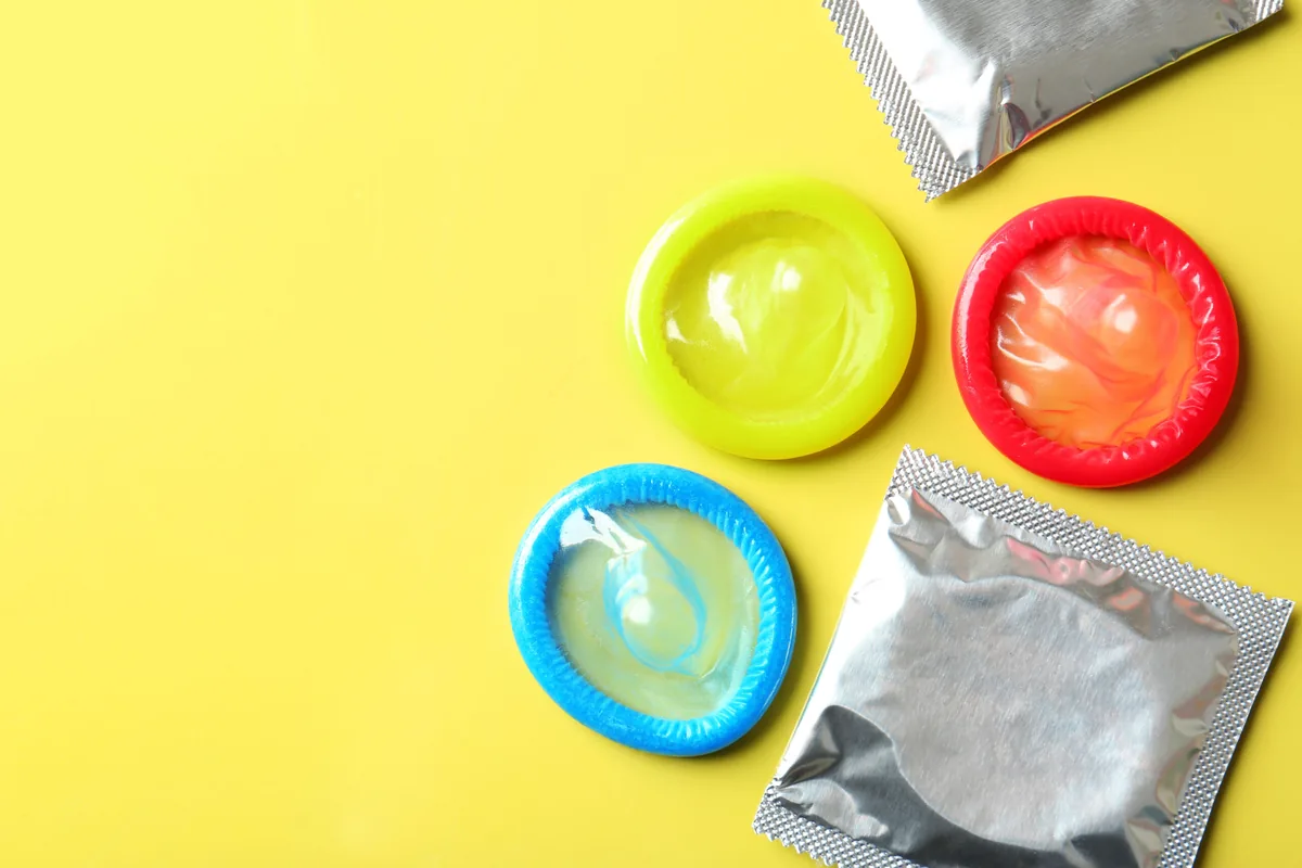Flat-lay photo of three unrolled condoms in green, orange and blue with two unbranded foil wrappers on a bright yellow background