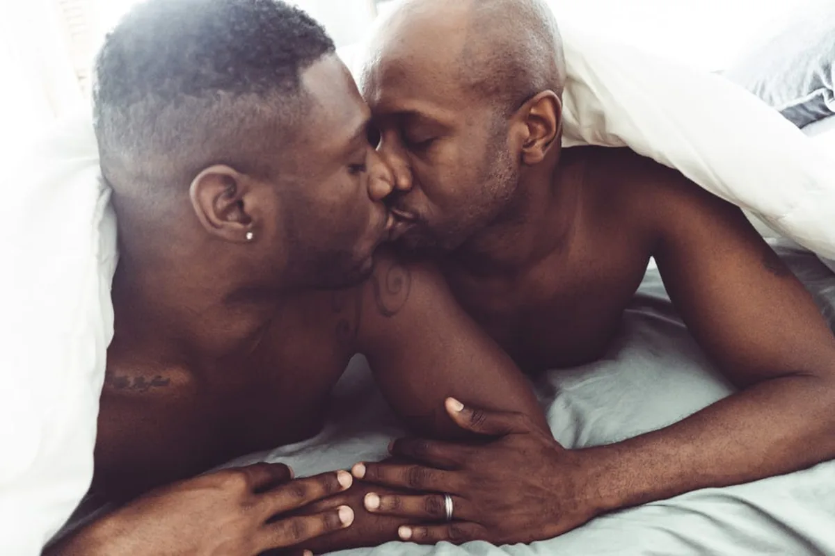 Two men kissing under a white duvet, holding hands