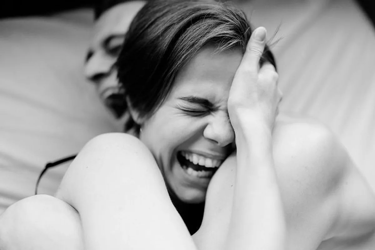 Black and white photograph of two people on a bed, one pressing a hand to their face while the other embraces them from behind