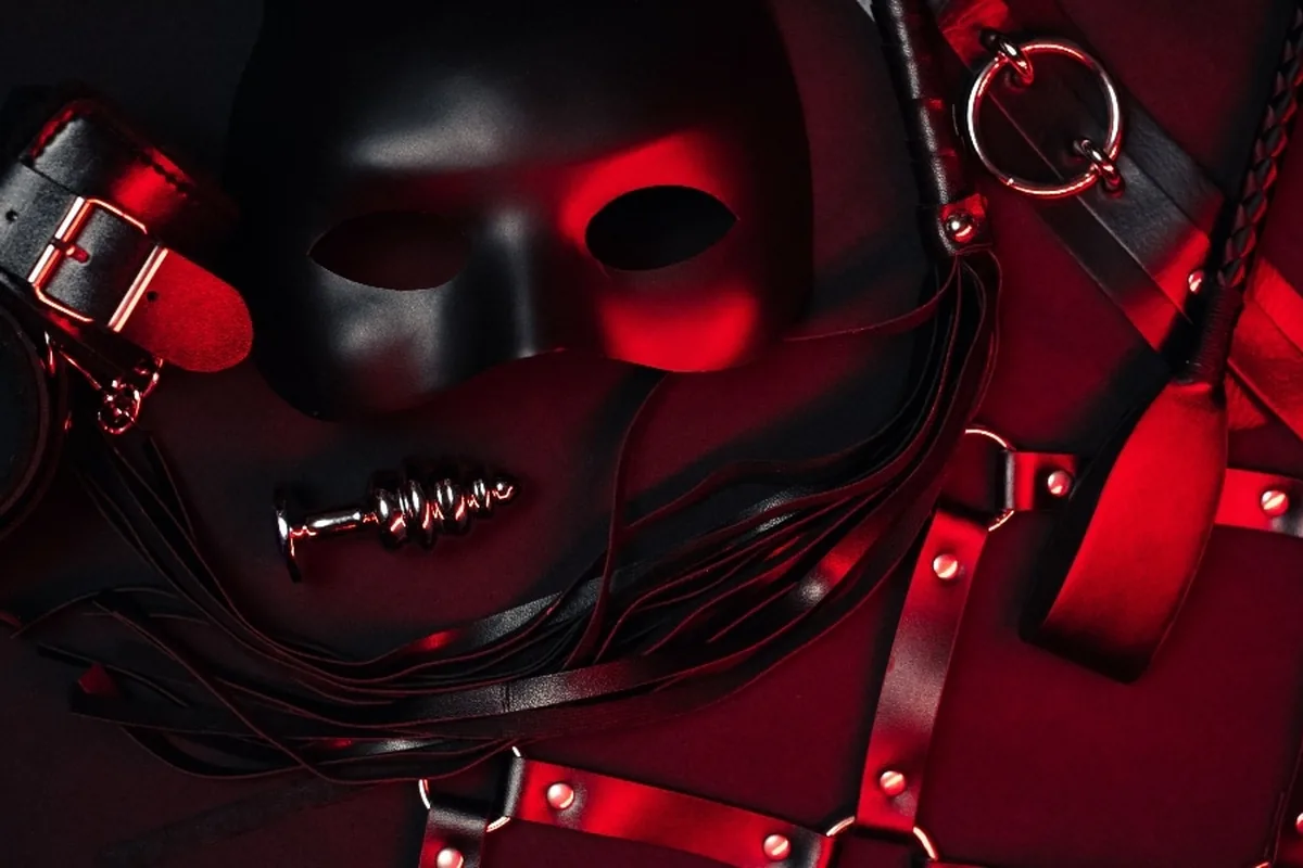 A black mask, leather cuffs, a harness and a flogger laid out on red fabric