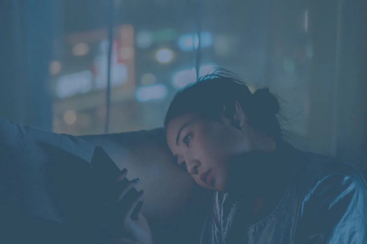 A woman lying on a couch in a dimly lit room at night, looking down at the glowing smartphone in her hands, with blurred city lights through the window behind her