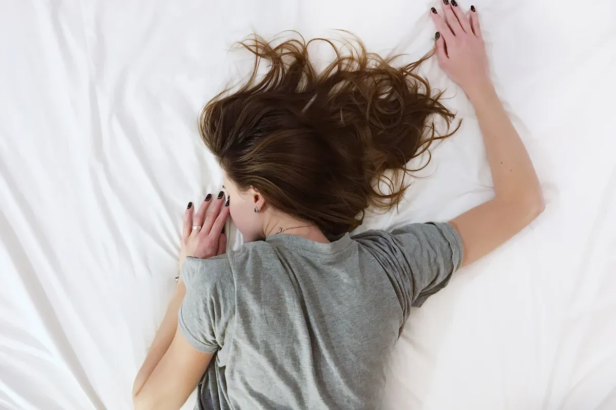 Overhead photo of a person with long brown hair lying face down on a white bed in a gray t-shirt, one arm stretched above their head