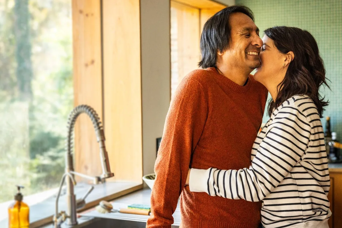 A woman in a striped long-sleeve top kissing the cheek of a smiling bearded man in an orange sweater as they embrace in a sunny home kitchen