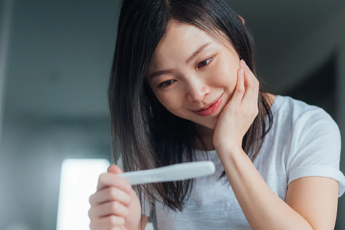 A smiling young woman in a white t-shirt holding a home pregnancy test in a softly lit room