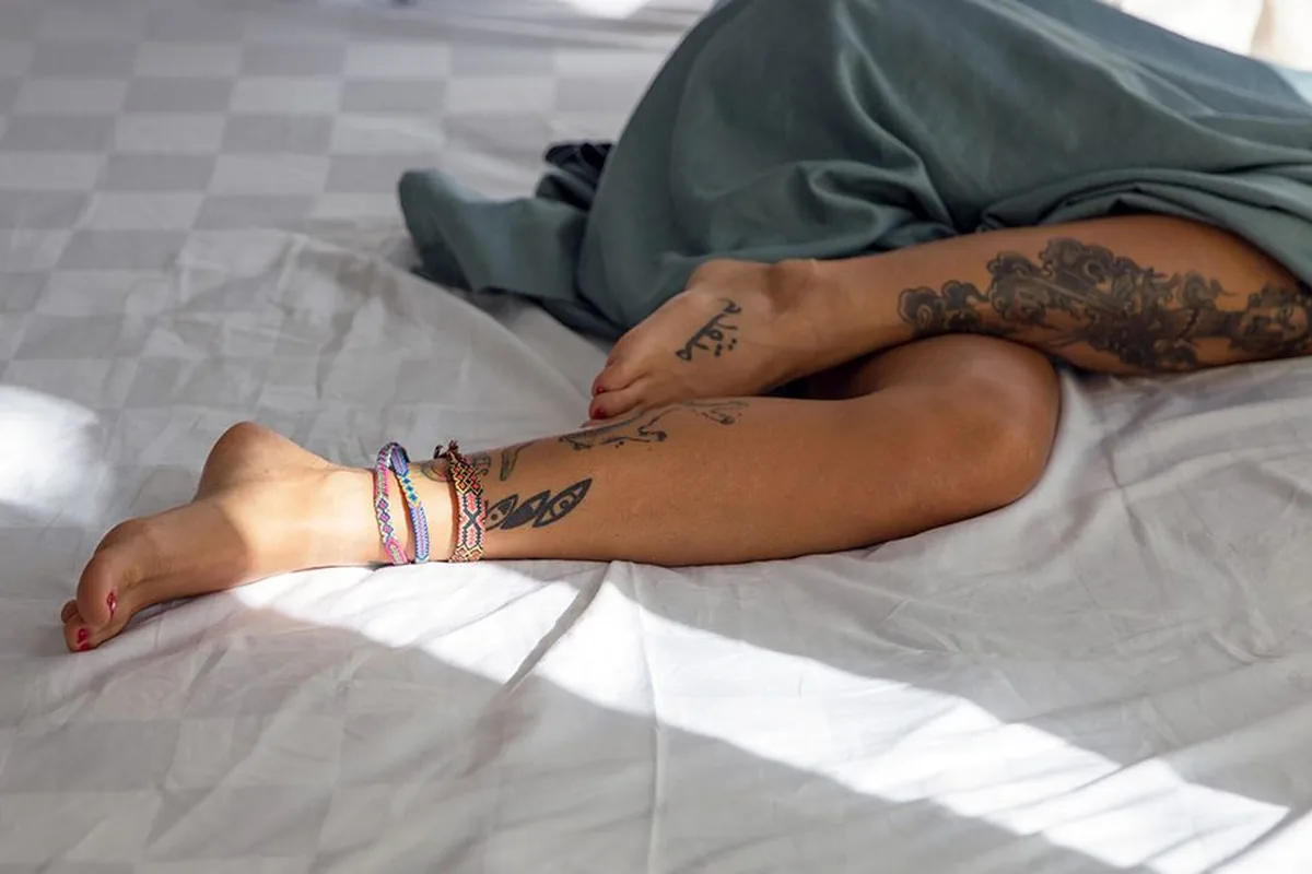 A person’s bare legs and feet resting on a white bed under a sage-green blanket, toenails painted red, in soft sunlight