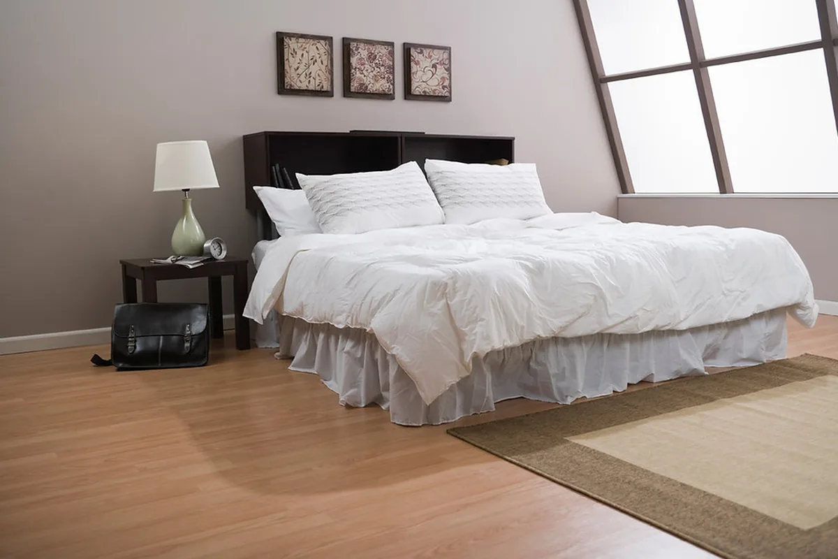 A neatly made bed with white pillows and bedding against a dark bookcase headboard, lit by a window in a quiet bedroom