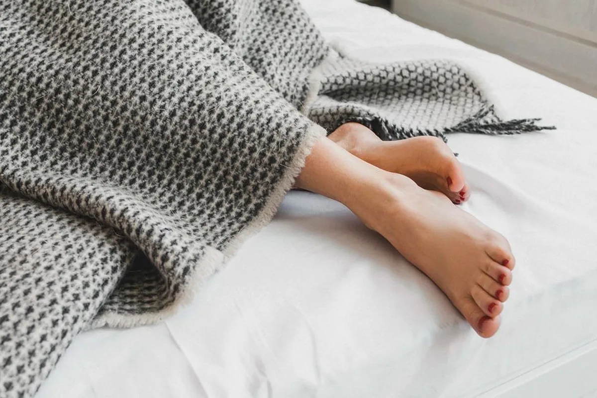 A close-up of a person’s bare feet and lower legs stretched out under a grey woven wool blanket on a bed with white sheets, toenails painted dark red