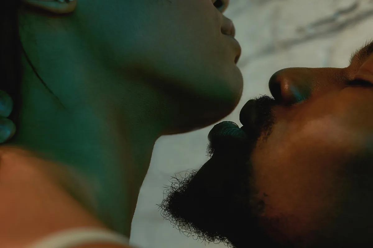 Two people lying close together on soft bedding, faces tilted toward each other just before a kiss