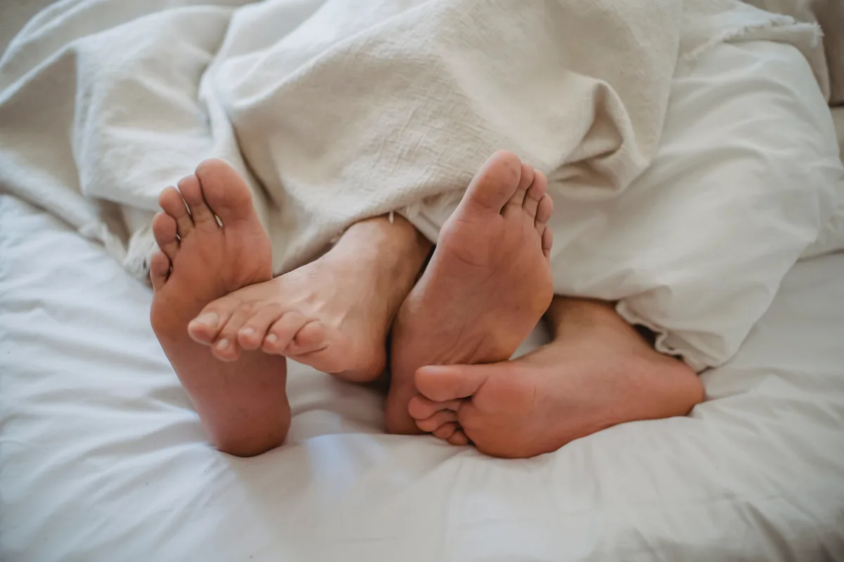 Two pairs of bare feet beneath a cream-colored blanket on a bed, a couple lying together under the covers