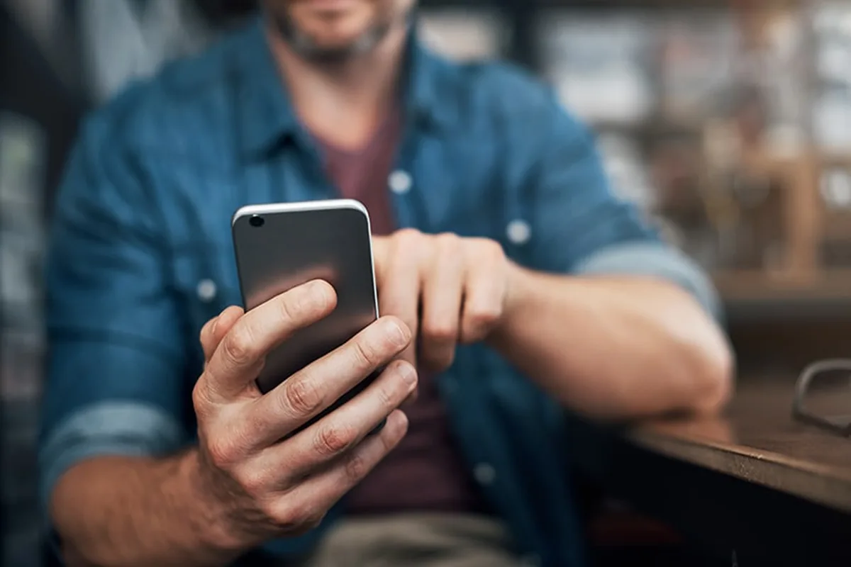 A man in a denim shirt seated in a warmly lit cafe, looking down at the phone in his hand as he checks information