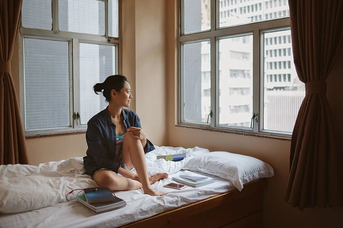 A young woman in an open denim shirt sitting cross-legged on a bed by large windows, gazing thoughtfully toward city high-rises