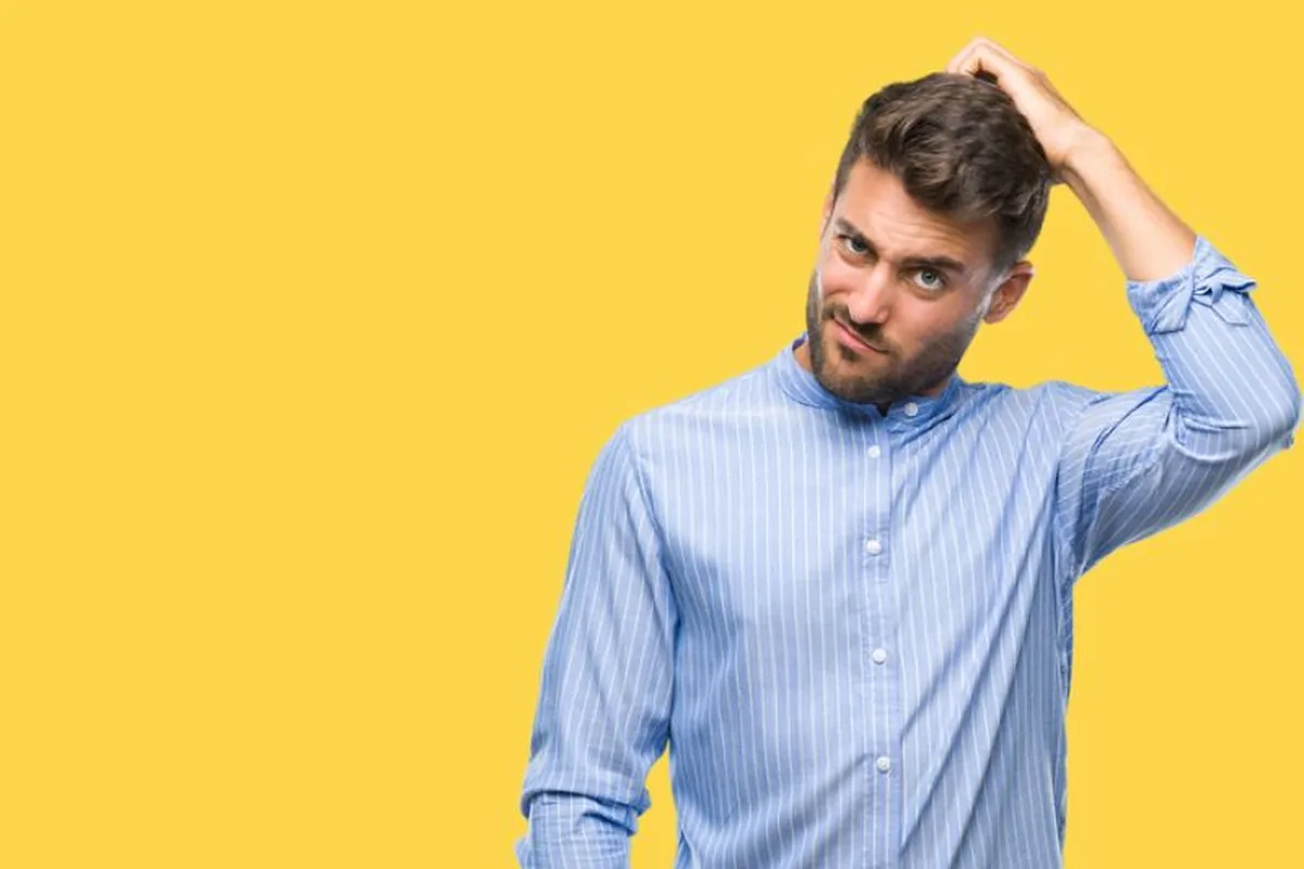 A puzzled young bearded man in a striped button-up shirt scratching his head against a plain yellow studio background