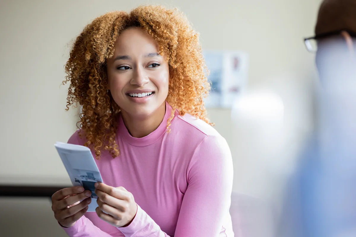 A smiling young woman with curly hair holding a brochure while talking with a healthcare provider in a consultation room
