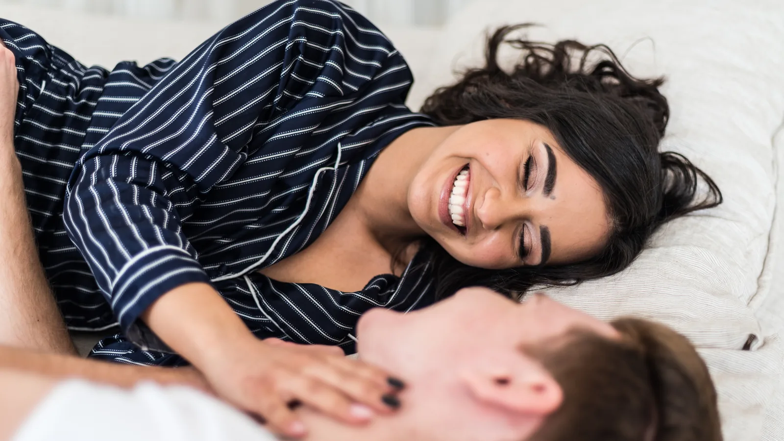 A couple in pajamas laughing while lying together on a white bed, her hand resting on his chest