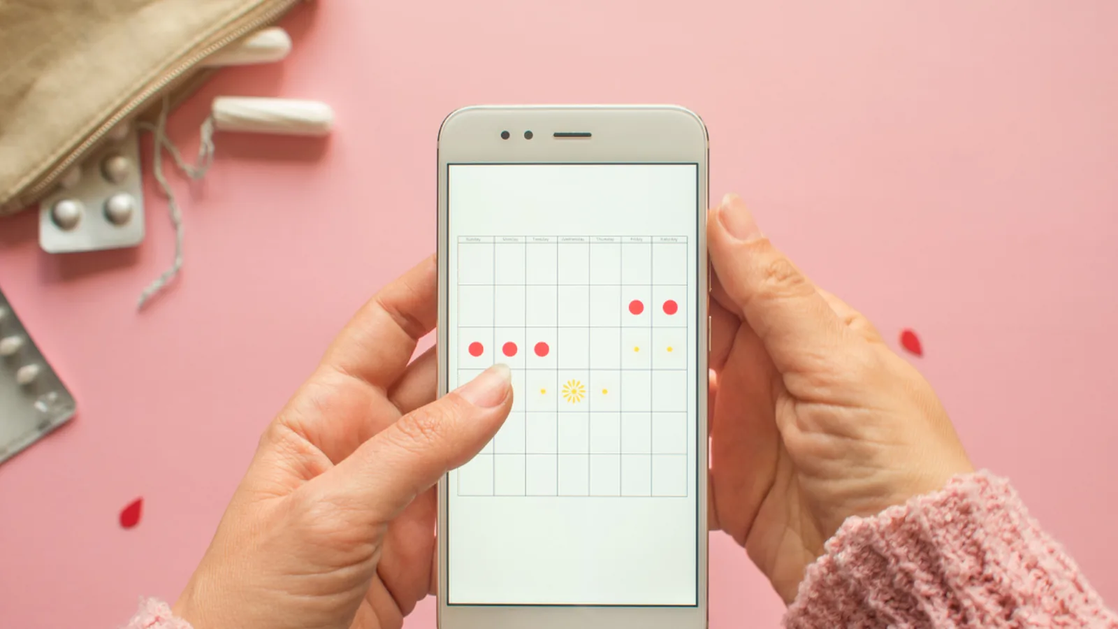 Hands holding a phone with a period-tracking calendar beside pills and tampons on a pink surface
