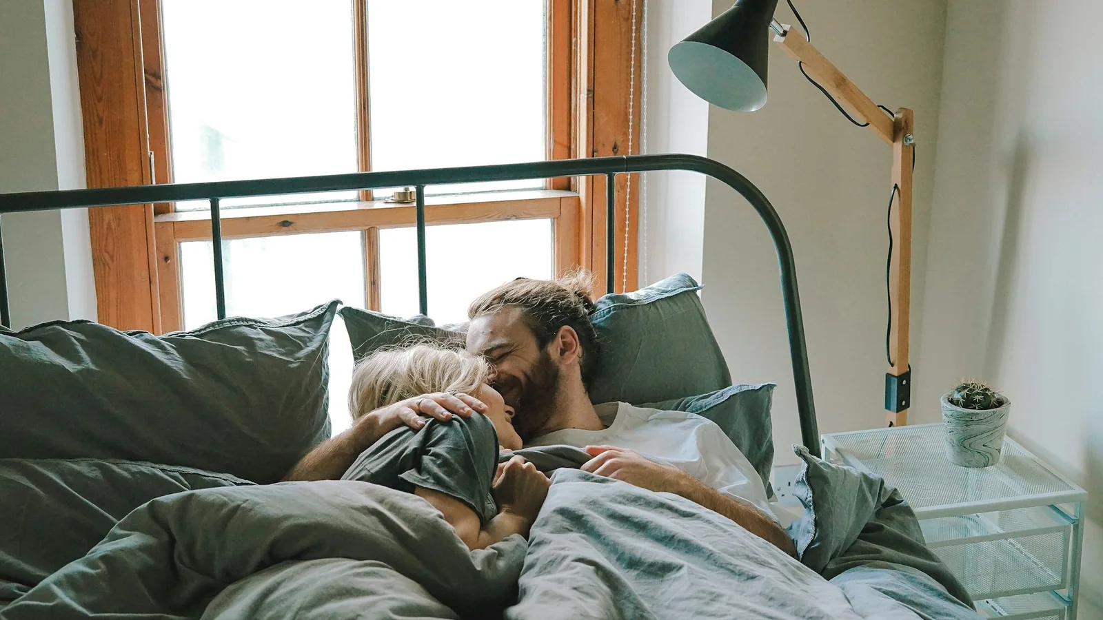 A clothed couple cuddling and embracing in bed in a bright bedroom with a green metal bed frame