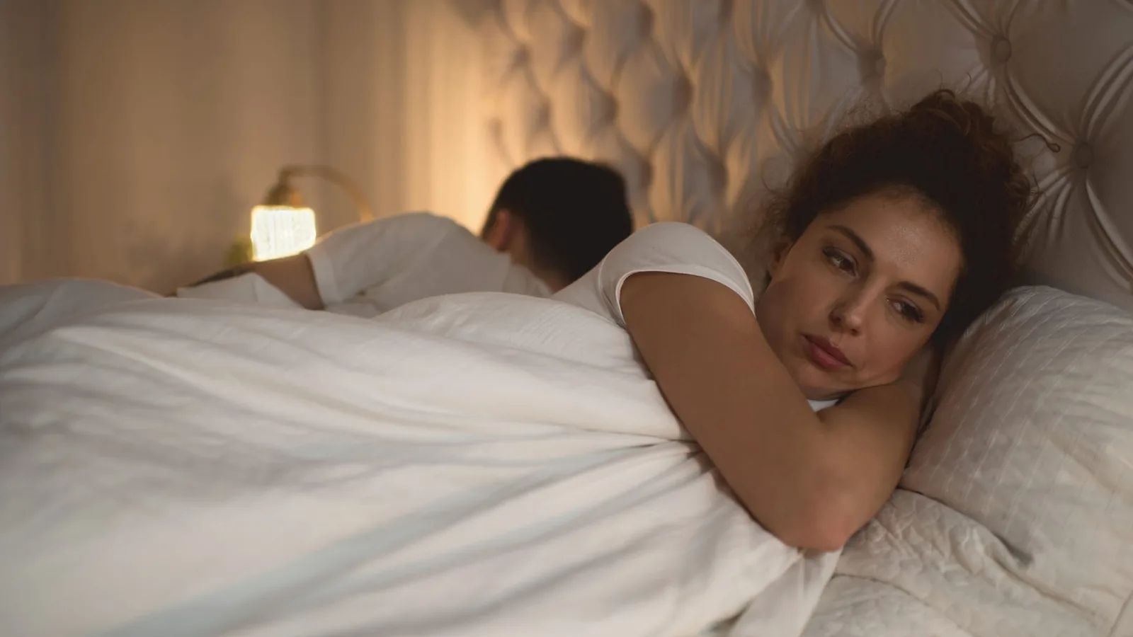 A woman lying awake with a troubled expression in bed at night while her partner sleeps turned away beside her, a bedside lamp glowing